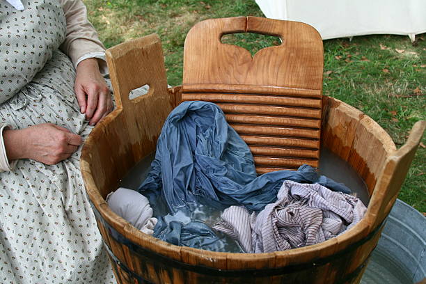 A wooden Colonial era washtub is seen filled with wet clothes and soapy water along with the wooden washboard used to scrub the clothes as the washer woman sits nearby.