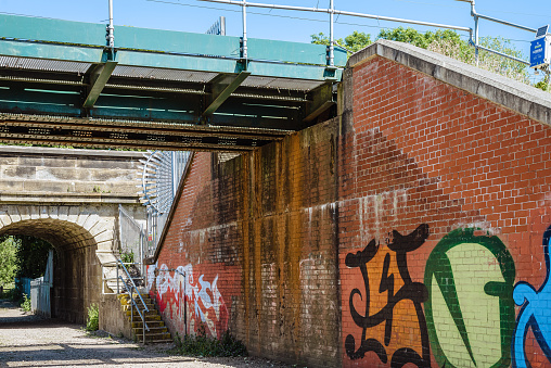 York, UK. May 29, 2020. Two railway bridges with one supporting wall covered in colourful graffiti. A tunnel leads to some trees and there is a blue sky above.