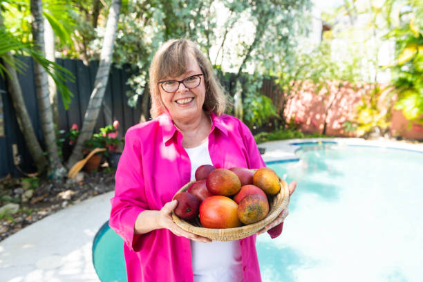 Vibrant Senior Woman in 60s Holding Homegrown Mangos in Backyard by the Pool