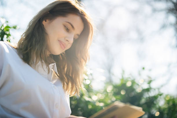 Smiling sly woman covering low face with open notebook with blank cover. Middle aged Caucasian lady reading notes outdoors, hiding face behind copybook and looking at camera. Keeping secret concept