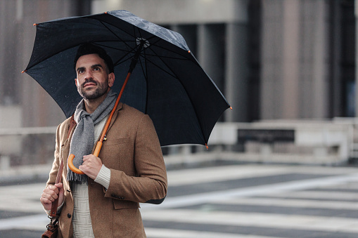 Cropped shot of a handsome young businessman holding an umbrella while standing in the rain outside during the day