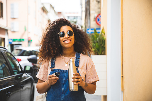 Portrait of a smiling afro hair woman holding a reusable water bottle waking in the city street in summertime.