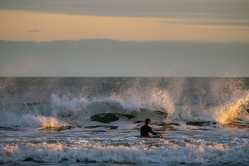 A wide shot of a young Caucasian man wearing a wetsuit, in the middle of surfing in the sea, the waves are crashing around him.