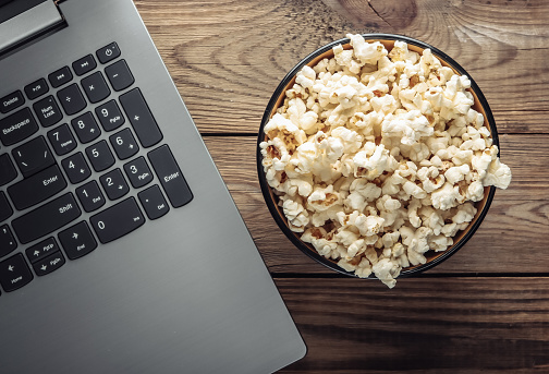 Laptop, bowl of popcorn on wooden table. Leisure and entertainment concept. Top view
