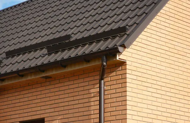 Photo of A close-up on a metal tiled rooftop with roof snow guards, snow stoppers, and a rain gutter with a downspout on a brick house.