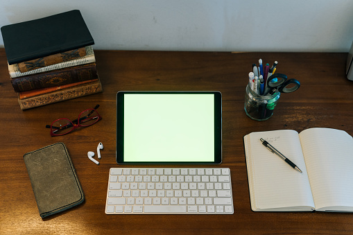 Tablet, keyboard, books, notepad, reading glasses, ear buds, and pens on workspace