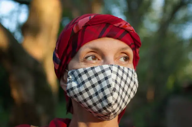 Photo of Mature woman wearing handmade textile face mask as accessory and protective element during sunny spring day in the park because of coronavirus pandemic