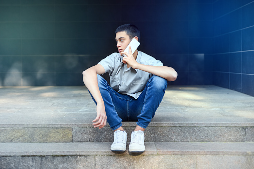 Young teenage boy chatting on his mobile phone outdoors sitting on city steps as he looks away to the side listening to the conversation with a thoughtful expression