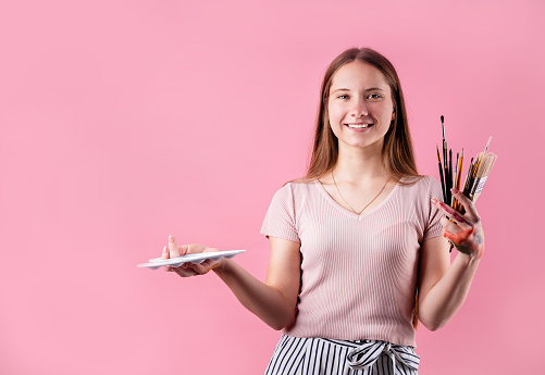 Female artist with a color palette and paintbrushes isolated on pink background with copy space