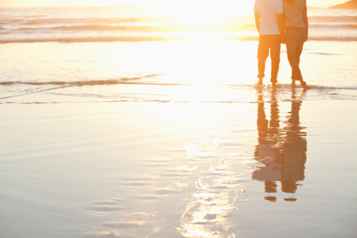 Romantic couple walking on sunset beach, enjoying evening light, relaxing on tropical summer vacation. Honeymoon. Love. Back view. Woman wearing orange maxi dress, man in linen pant and white shirt.