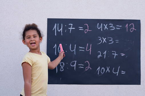 An excited pupil girl studying arithmetic math, learning division, and multiplication. The elementary school child pointing with pink chalk, showing math exercises written on a black chalkboard.