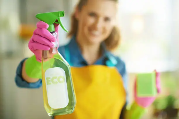 Closeup on happy woman in orange apron and pink rubber gloves with green sponge showing spray bottle of eco friendly cleaning supplies at home in sunny day.