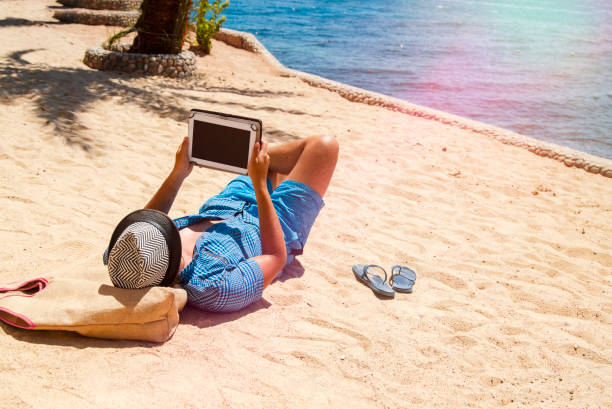 jeune homme recherchant un site web de voyage pour des hôtels ou des resorts sur les vacances d’été - reading beach e reader men photos et images de collection