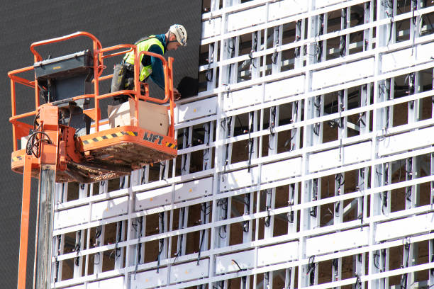 Construction worker in crane basket installing sheets cladding for covering structure on a building facade wall stock photo