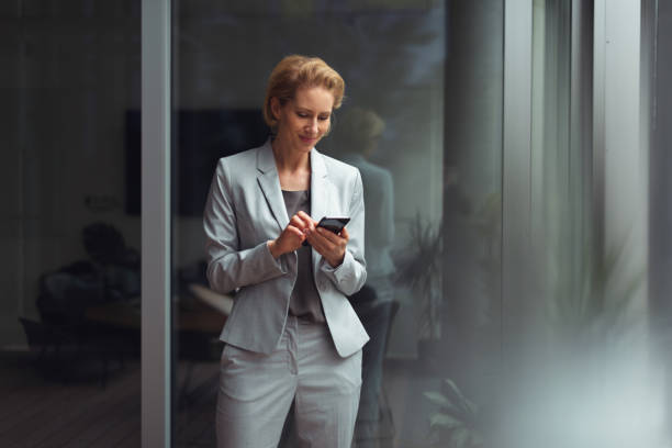 German businesswoman 40-year-old satisfied caucasian businesswoman is standing in office and using smartphone. georgijevic frankfurt stock pictures, royalty-free photos & images
