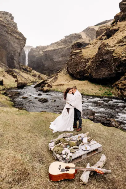 Photo of Destination Iceland wedding, near Kvernufoss waterfall. A wedding couple stands under a plaid near a mountain river. The groom hugs bride. They built an impromptu wedding table with decor and guitar