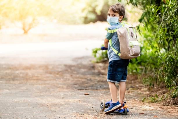 little boy riding a push scooter carrying a back pack wearing a protective face mask - little boys preschooler back to school backpack imagens e fotografias de stock