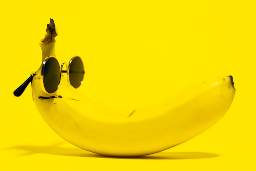 Close-up of yellow banana wearing round sunglasses lying on background of yellow color.
