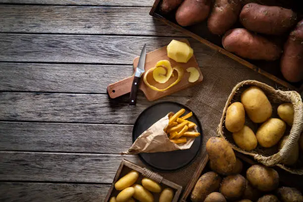 French fries potatoes on paper cone on a rustic wooden board table with raw potatoes around of different colors and sizes shapes