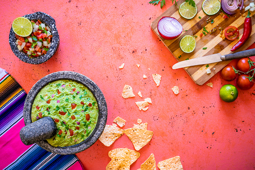 Guacamole Mexican avocado recipe with pico de gallo sauce in stone molcajete tortilla chips and ingredients on coral pink background