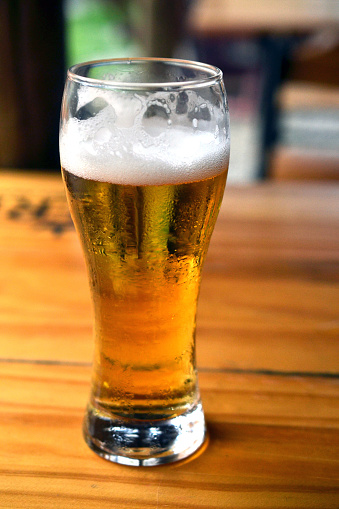 A golden pint of beer with foam at the top of the glass.  The glass of beer is placed on a hardwood surface with liquid drops of beer surrounding the glass on the table.