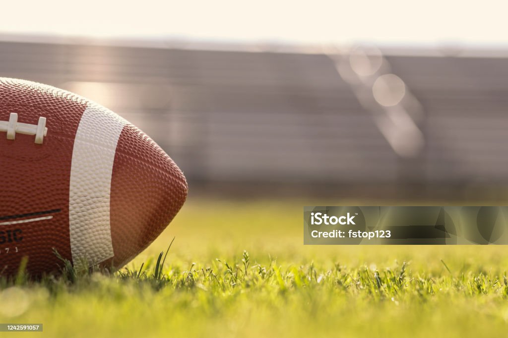 American football on stadium field at school campus. Football on grass stadium on college or high school campus. Bleachers background. No people.  Daytime. American Football - Sport Stock Photo