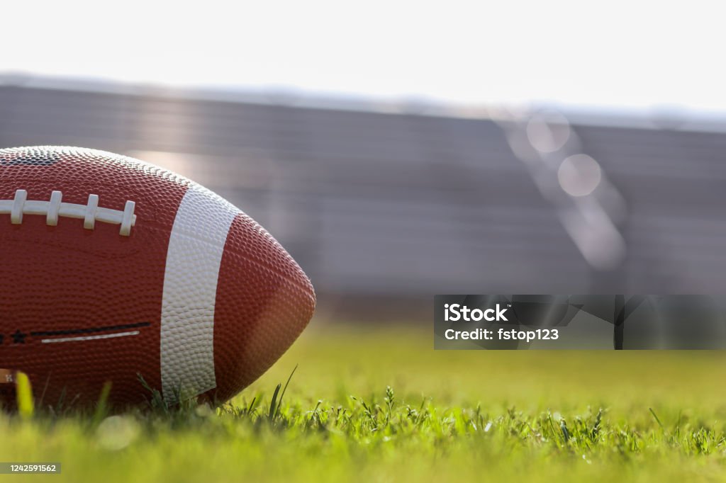 American football on stadium field at school campus. Football on grass stadium on college or high school campus. Bleachers background. No people.  Daytime. American Football - Ball Stock Photo