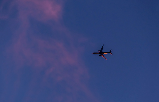 Silhouette of an unidentified commercial passenger jet plane glows red in the sunset over New Jersey as it flies towards wispy pink clouds.  Metaphor for problems with the aviation industry due to pandemic, economic downturn and climate change.