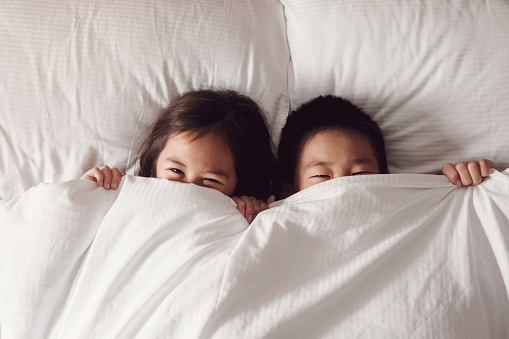 Happy young Asian brother and sister covering their faces with blanket and peeking out cheerfully in the morning