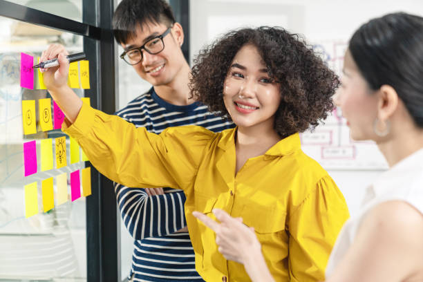 istock Asian creative woman smiling and meeting at office use post it notes to share idea and planning project. Group of employee brainstorming idea on glass wall and sticky. Brainstorming concept. 1242740095