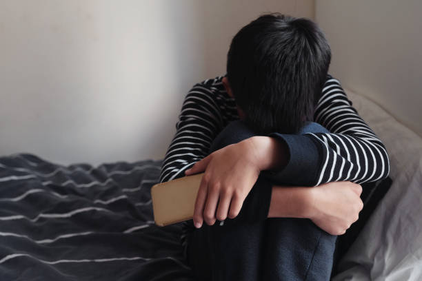 giovane ragazzo adolescente preteen asiatico che abbraccia il ginocchio nella sua camera da letto con lo smartphone, cyber bullismo nel bambino, salute mentale dei bambini depressa - cyberbullismo foto e immagini stock