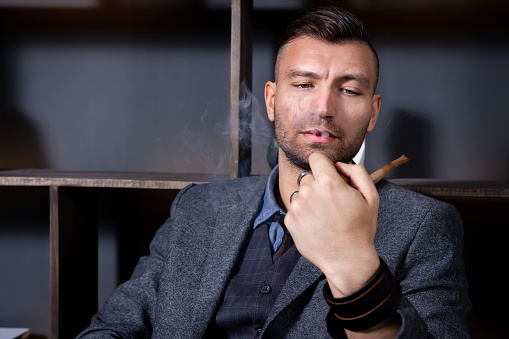 Portrait of elegant man wich sits in armchair and smokes in his workoffice. Businessman relaxes after hard day.