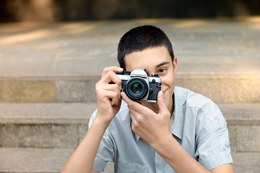 Young teenage boy photographing the viewer with a friendly smile as he focuses his shot while seated outdoors on urban steps