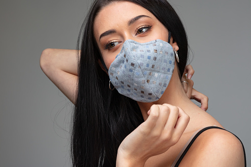 Young woman with long dark hair wearing a stylish cloth face mask against Covid-19 or coronavirus infection in a concept of the new fashion reality over grey