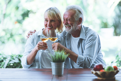 Senior couple celebrating retirement outdoor.  White man and woman. Holding white wine glasses with tropical garden in background. Toasting and laughing.