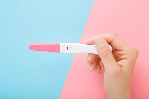 Joven mujer sosteniendo la prueba de embarazo con dos rayas sobre fondo de mesa azul rosa claro. Color pastel. Dos lados. Resultado positivo. Closeup. Disparo de punto de vista. Vista de arriba hacia abajo. photo