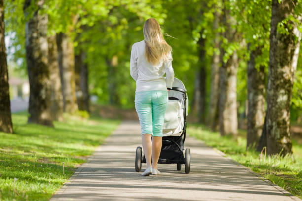 una joven madre empujando cochecito blanco y caminando lentamente por el callejón de los árboles en un cálido y soleado día de primavera. pasar tiempo con el bebé. disfrutando de un paseo. vista trasera. - baby mother summer park fotografías e imágenes de stock