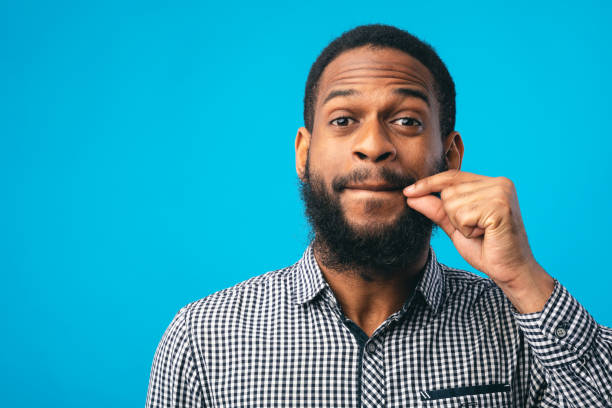 Funny black man making zipping lips gesture with hand I Won't Say A Word. Black bearded guy making zipping lips gesture with hand, showing he will keep a secret, blue studio airtight photos stock pictures, royalty-free photos & images