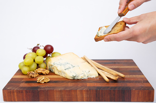Human hand spreading cheese on toast. Blue cheese, smoked cheese sticks, grape and walnut on cutting board. Cheese snack concept