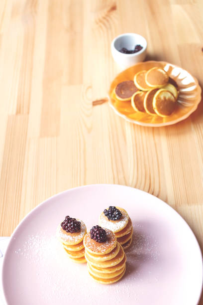 Trendy tiny pancakes cereal meal on pink plate. Baked pancakes for breakfast on wooden background. Fresh berries for decoration in background.
