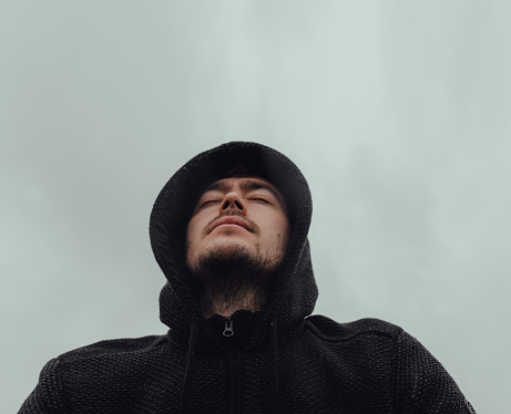 From below calm bearded male in black hoodie closing eyes and breathing deeply against cloudy gray sky in nature