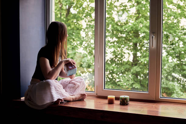 Young brunette woman, wearing black top and light pale pink yoga pants, sitting on windowsill with colorful aroma candles, looking through at green trees in park, thinking, meditating.