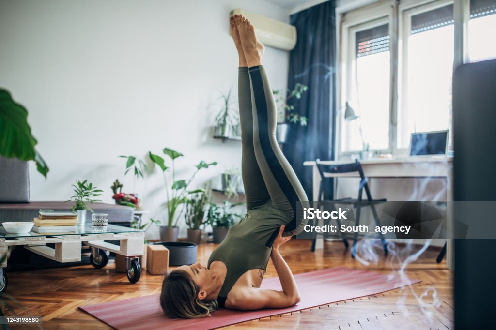 Woman lying on upper back and holding her lower back in balance One beautiful young woman doing yoga on exercise mat in her living room, lying on upper back and holding her lower back in balance. Women Stock Photo