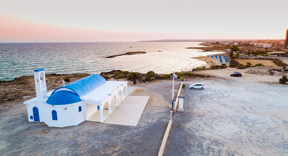 Aerial view of coastline sunset and traditional landmark white washed chapel with blue doors at Agia Thekla beach, Ayia Napa, Famagusta, Cyprus from above. Bird's eye view of tourist attraction golden sand bay, church, sunbeds in Ammochostos.