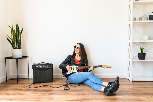 A young beautiful woman is playing an electric guitar using a combo amplifier, she is in a black leather jacket and in sunglasses sits on the floor at home