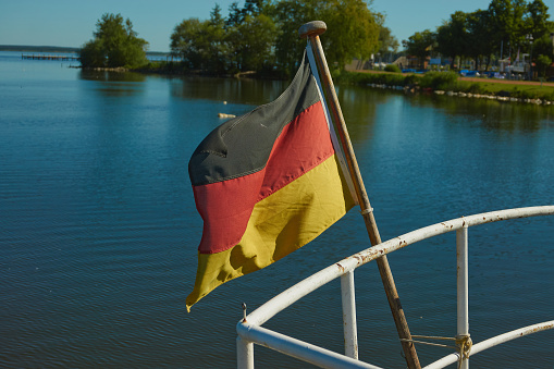 Black-red-golden flag of Germany on the stern of a ship on a blue lake with green bushes on the shore