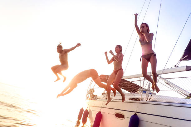 Young millenial friends jumping from sailboat at sea ocean trip - Guys and girls having summer fun together at sail boat party day - Luxury excursion concept on bright vivid filter with soft focus Young millenial friends jumping from sailboat at sea ocean trip - Guys and girls having summer fun together at sail boat party day - Luxury excursion concept on bright vivid filter with soft focus catamaran sailing boats stock pictures, royalty-free photos & images