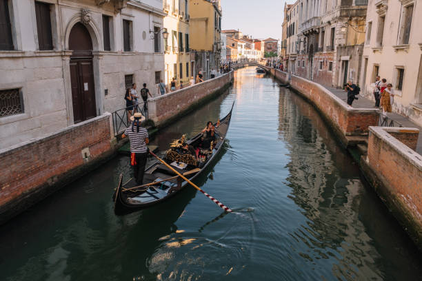 gondolier drives a gondola with two tourists on a canal in venice, italy. they have a date. woman playing the violin. tourists on the shore smile at them and take pictures. - couple performer people venice italy imagens e fotografias de stock