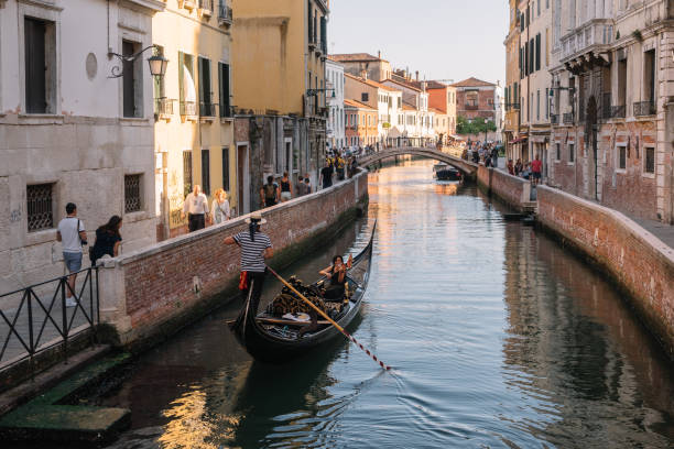 gondolier drives a gondola with two tourists on a canal in venice, italy. woman playing the violin. they have a date. tourists on the shore smile at them and take pictures. - couple performer people venice italy imagens e fotografias de stock