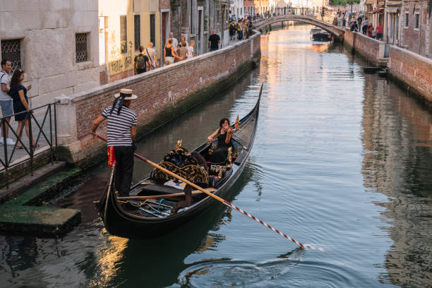 gondolier conduce una góndola con dos turistas en un canal en venecia, italia. tienen una cita. mujer tocando el violín. los turistas en la orilla sonríen y toman fotos. - couple performer people venice italy fotografías e imágenes de stock